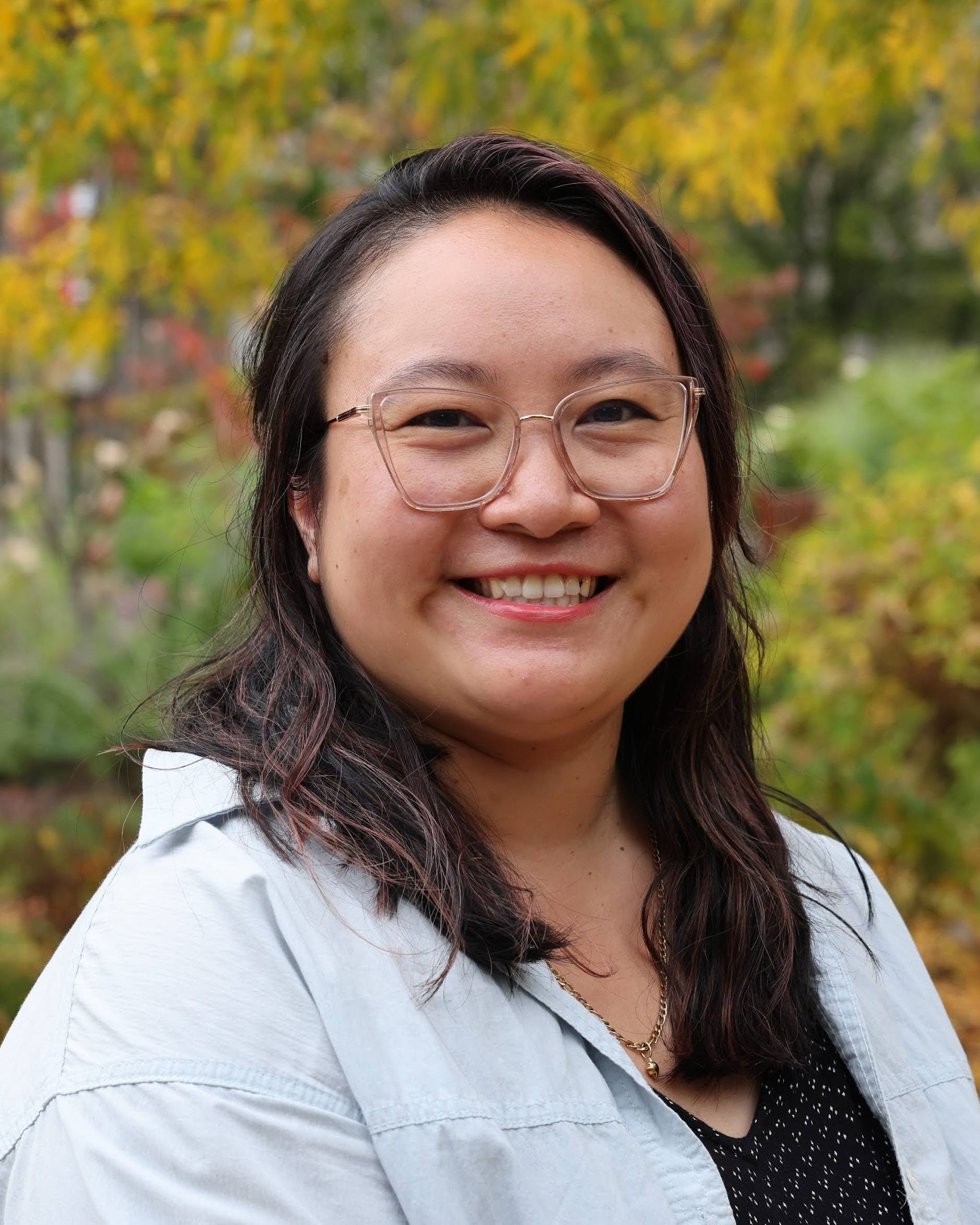 Image of Joy, a smiling Chinese-American woman in her 30's with pink glasses standing in front of orange and yellow fall leaves.