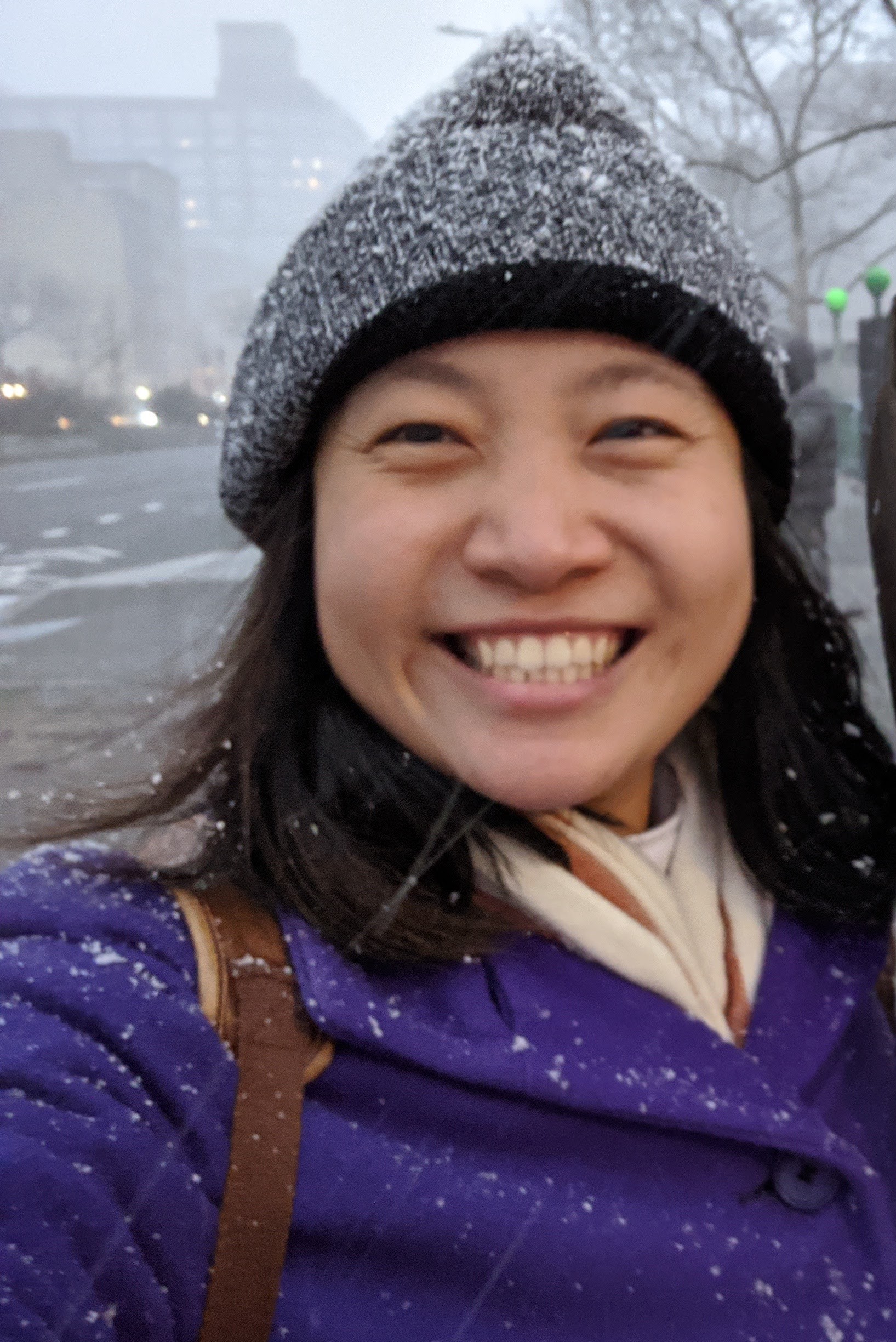 Image of Joy, a smiling Chinese-American woman in her late 20's wearing a grey beanie, beige scarf, and purple jacket in the snow.